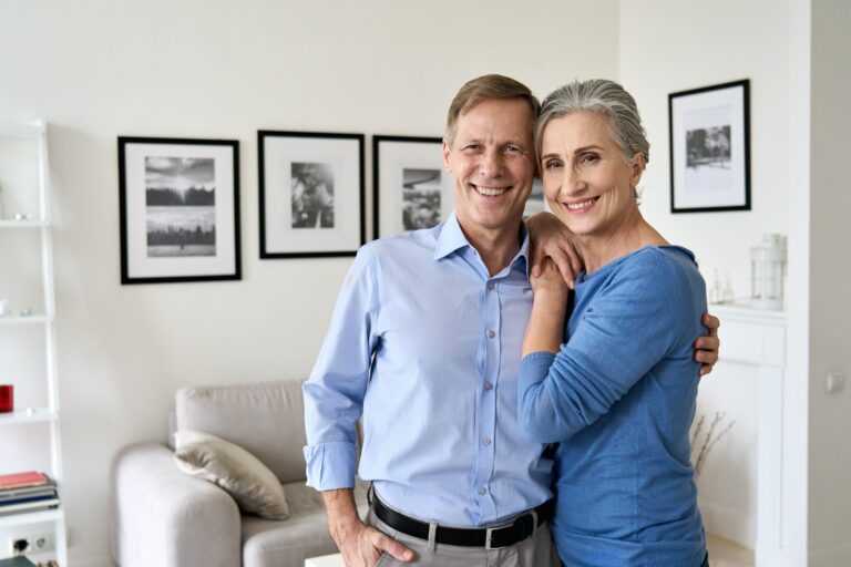 Happy older couple hugging, looking at camera, sitting on couch in apartment.
